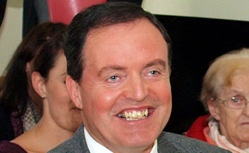 Ein Mann lächelt breit mit sichtbaren Zähnen und grauem Anzug vor zwei Frauen.