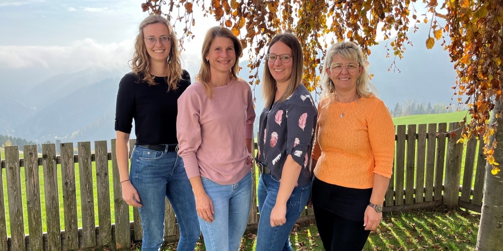 Vier Frauen in lässiger Kleidung stehen draußen in einem Feld und lächeln. Hinter ihnen befinden sich ein Holzzaun und Bäume. Der Hintergrund zeigt ein Grasfeld und Berge.