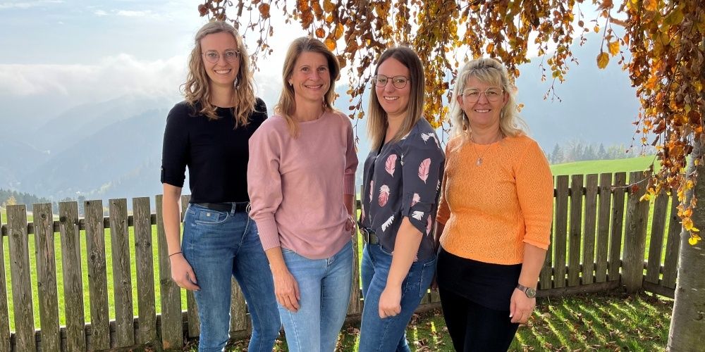 Vier Frauen in lässiger Kleidung stehen draußen in einem Feld und lächeln. Hinter ihnen befinden sich ein Holzzaun und Bäume. Der Hintergrund zeigt ein Grasfeld und Berge.