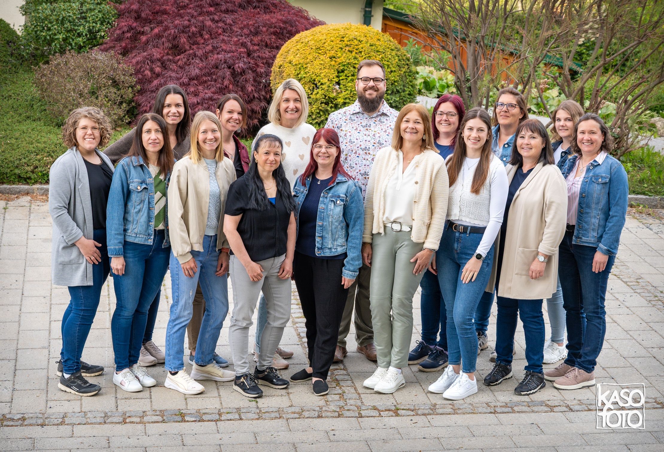 Eine Gruppe von Frauen und einem Mann steht auf einem gepflasterten Weg und trägt alle Freizeitkleidung und Sneaker. Sie posieren für ein Foto in einem Garten mit verschiedenen Pflanzen und Bäumen im Hintergrund.