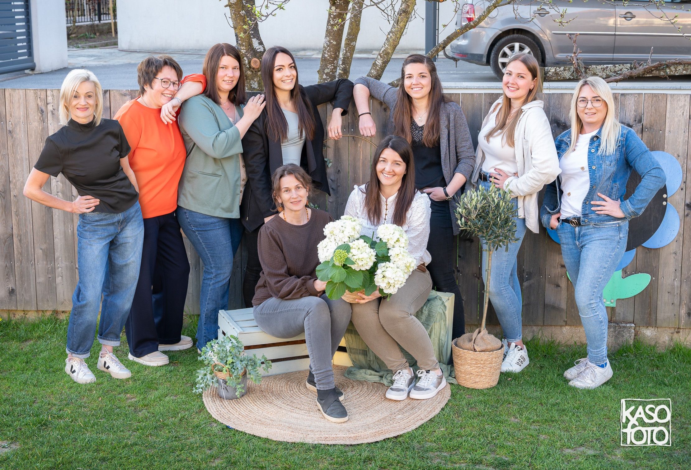 Eine Gruppe von Frauen und Männern posiert für ein Foto im Garten mit Bäumen, einem Zaun und Pflanzen. Eine Frau hält Blumen.