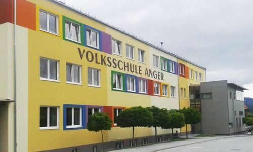 Das Volksschule Anger Gebäude ist gelb gestrichen mit farbenfrohen Fenstern, die den Namen deutlich anzeigen. Das Gebäude befindet sich neben einem größeren Gebäude mit Glasfenstern.