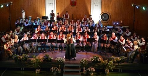 Ein großes Orchester spielt auf der Bühne mit verschiedenen Instrumenten, umgeben von Blumen und Holzwänden, wobei Scheinwerfer die Szene beleuchten.