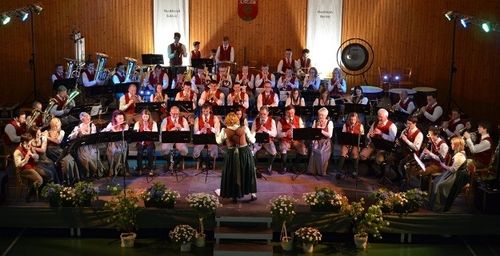 Ein großes Orchester spielt auf der Bühne mit verschiedenen Instrumenten, umgeben von Blumen und Holzwänden, wobei Scheinwerfer die Szene beleuchten.