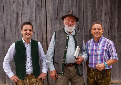 Drei Männer in traditioneller deutscher Kleidung stehen vor einer Holzwand. Der Mann in der Mitte trägt einen Hut und hält ein Buch. Der Mann auf der linken Seite trägt ein weißes Hemd, während der Mann auf der rechten Seite ein kariertes Hemd trägt.