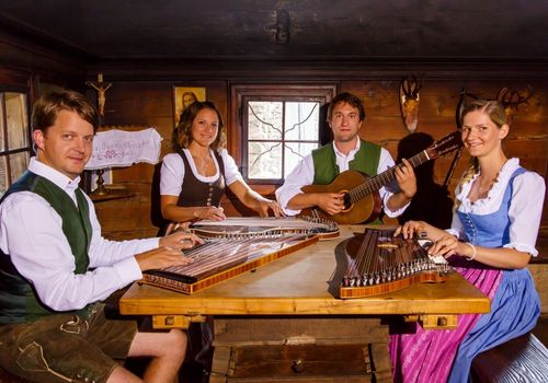Vier Personen in traditioneller Kleidung sitzen um einen Holztisch herum und spielen verschiedene Saiteninstrumente, einschließlich Gitarren und Zithern, in einem hölzernen Raum mit einem Fenster.
