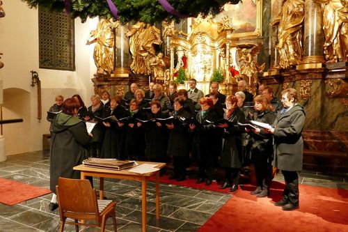 Ein Chor, in Schwarz gekleidet, steht vor einem goldenen Altar in einer Kirche und singt mit geschlossenen Augen. Der Chor hat einen roten Teppich vor sich.