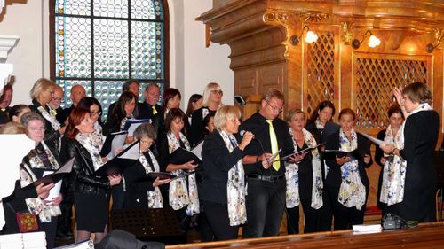 Ein Chor singt, wobei die Mitglieder Songbücher und ein Mikrofon halten. Der Ort ist mit einem Buntglasfenster und einer Holzstruktur ausgestattet.