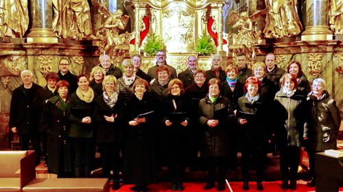 Ein Chor älterer Personen, sowohl Männer als auch Frauen, posiert vor einem Kirchenaltar für ein Foto. Sie halten Notenblätter in den Händen und tragen Winterkleidung.