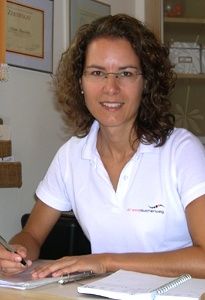 Eine Frau in einem weißen Poloshirt sitzt an einem Schreibtisch, schreibt mit einem Stift, mit Papieren und einem Notizbuch vor ihr und einem gerahmten Bild an der Wand hinter ihr.