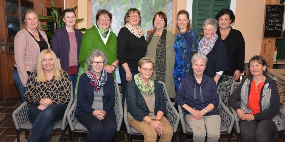Eine Gruppe von Frauen posiert für ein Foto in einem Raum. Einige sitzen auf Stühlen, während andere stehen. Alle lächeln für die Kamera.