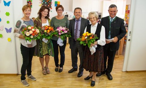 Sechs Menschen stehen in einem Raum und halten Blumensträuße. Drei Frauen tragen Kleider und drei Männer tragen Anzüge. Hinter ihnen ist eine Küche mit Schränken und einer Mikrowelle.