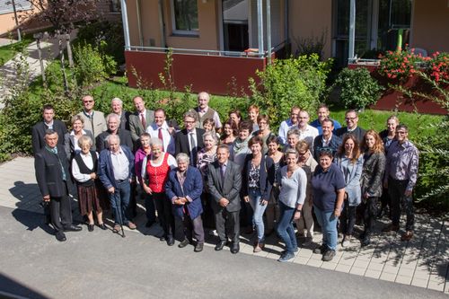 Ein Gruppenfoto von formell gekleideten Personen vor einem Gebäude mit Pflanzen und Rasen im Hintergrund.