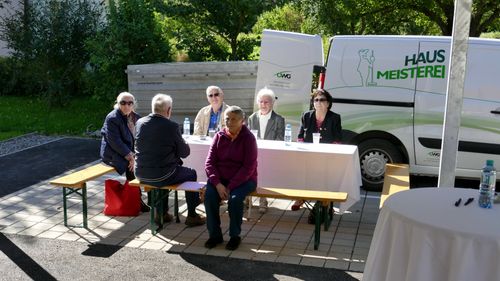 Eine Gruppe älterer Personen sitzt um einen Tisch herum draußen, mit einem Van mit der Aufschrift 'OWG' und 'MW' dahinter und Bäumen im Hintergrund.