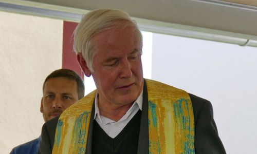 Ein älterer Mann mit weißen Haaren, der eine schwarze und gelbe Stola trägt, steht und schaut nach unten. Ein anderer Mann in einem blauen Hemd steht hinter ihm.