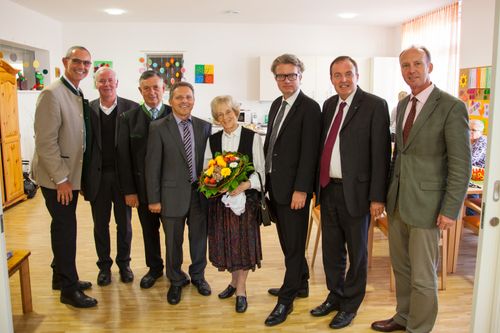 Eine Gruppe von formell gekleideten Männern und einer Frau mit Blumen steht in einem Raum. Sie lächeln für ein Foto.