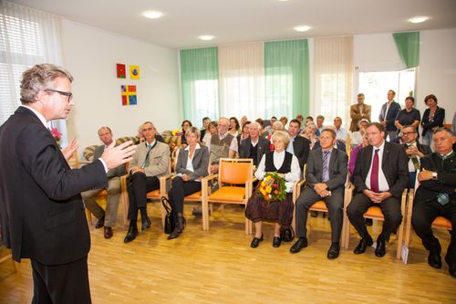 Eine formelle Versammlung in einem Innenraum, mit einem Sprecher vor einem sitzenden Publikum. Die Frau in der Mitte hält einen Blumenstrauß. Der Raum ist gut beleuchtet mit Kunstwerken an der Wand.
