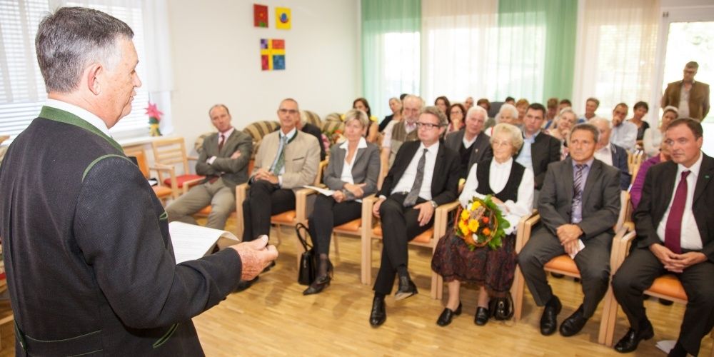 Eine Gruppe von Menschen ist in einem Raum versammelt, möglicherweise für eine Besprechung. Einige von ihnen sitzen auf Stühlen, während andere stehen. Der Mann, der vor ihnen steht, hält ein Papier in der Hand und scheint den Leuten etwas zu sagen. Der Raum hat einen Holzboden, eine weiße Wand mit Vorhängen und Gemälden.