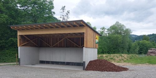 Ein kleines Holz- und Betongebäude mit einem Metalldach. Vor ihm befindet sich ein Haufen braunen Materials. Bäume und Büsche umgeben das Gebiet.