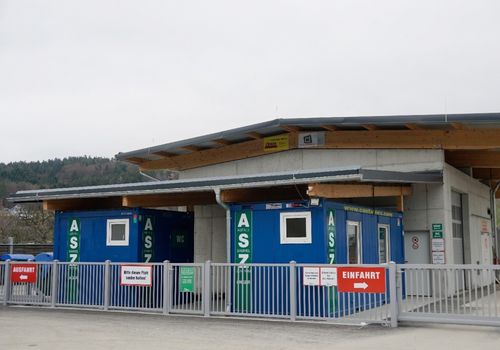 Ein blauer und grüner Container mit dem Wort ASZ und WC darauf befindet sich hinter einem Zaun. Ein Schild am Zaun sagt Bitte diesen Platz sauber halten. Ein weiteres Schild sagt Einfahrt mit einem Pfeil nach rechts.