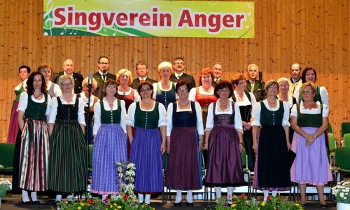 Eine Gruppe von Menschen in traditioneller Kleidung steht auf der Bühne mit einem Banner im Hintergrund. Einige von ihnen halten Mikrofone.