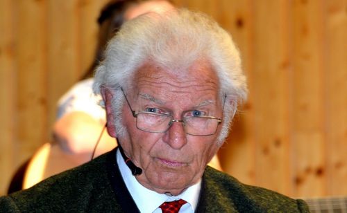 Ein älterer Mann mit weißen Haaren, einer Brille und einer roten Krawatte sitzt in einem Raum und trägt ein Mikrofon.
