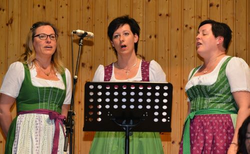 Drei Frauen in traditionellen Kleidern singen in Mikrofone vor einer Holzwand, eine steht hinter einem Notenständer.