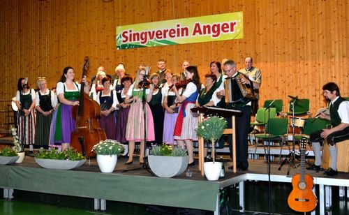 Eine Gruppe von Musikern in traditioneller Kleidung tritt auf der Bühne auf, begleitet von einem Schild, das 'Singverein Anger' steht. Dazu gehören Geiger, ein Akkordeonspieler und ein Cellist, mit Topfpflanzen und Stühlen im Hintergrund.