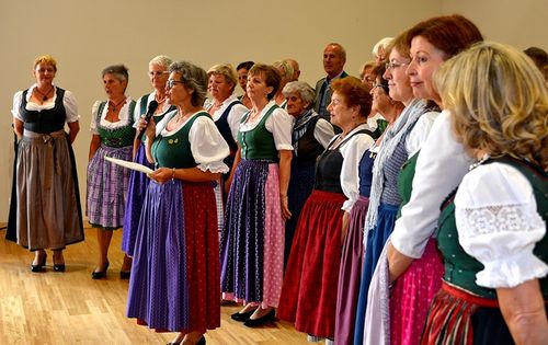 Eine Gruppe von Frauen in traditioneller Kleidung, möglicherweise beim Singen, wobei eine eine Mikrofon und eine andere Papiere hält. Ein Mann steht unter ihnen. Sie sind drinnen auf einem Holzboden.