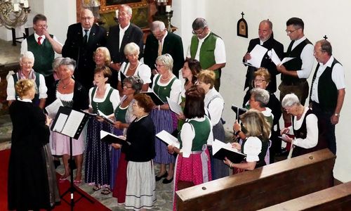 Eine Gruppe von Menschen in traditioneller Kleidung, sowohl Männer als auch Frauen, sind drinnen versammelt und singen gemeinsam. Sie halten Notenblätter in den Händen und scheinen eine Aufführung darzubieten.
