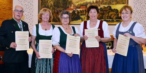 Drei Frauen in traditioneller Kleidung, lächelnd und Zertifikate in der Hand, stehen vor einem malerischen Gemälde. Sie scheinen einen Meilenstein zu feiern.