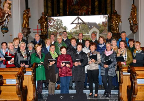 Eine Gruppe von Menschen, in Winterkleidung gekleidet, steht vor einer Kirche und hält Notenblätter in den Händen. Sie sind für eine Choraufführung versammelt, mit Statuen und einem großen Fenster im Hintergrund.