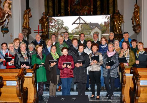 Eine Gruppe von Menschen, in Winterkleidung gekleidet, steht vor einer Kirche und hält Notenblätter in den Händen. Sie sind für eine Choraufführung versammelt, mit Statuen und einem großen Fenster im Hintergrund.