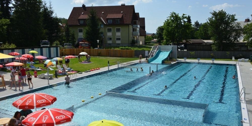 Ein Freibad mit Menschen, die schwimmen, rutschen und auf der Wiese unter Sonnenschirmen faulenzen. Es gibt Liegestühle und ein Gebäude im Hintergrund.