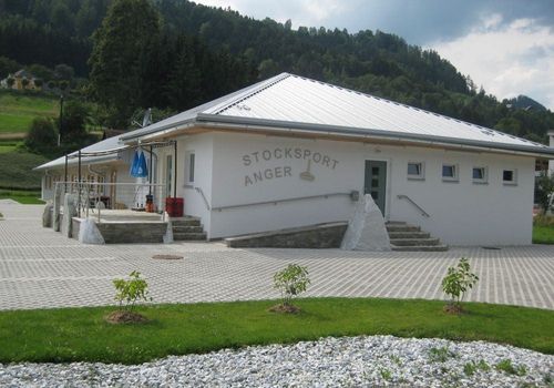 Das Stocksport Anger-Gebäude, weiß, mit Stufen und Geländern, hat ein Metalldach und ist von Kies und Pflanzen umgeben. Es hat mehrere Fenster und ein blaues Tuch, das draußen hängt.