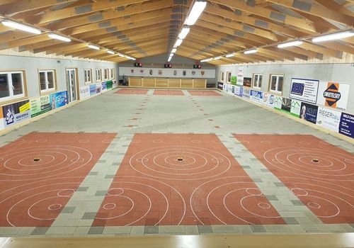 Eine leere Indoor-Sporteinrichtung mit gefliestem Boden, Holzbalken und beleuchteten Lampen. Es weist rote und weiße Markierungen auf dem Boden auf.