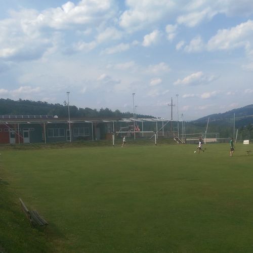Ein Fußballfeld mit einigen Spielern unter einem teilweise bewölkten Himmel. Ein Gebäude ist im Hintergrund mit Bergen dahinter.