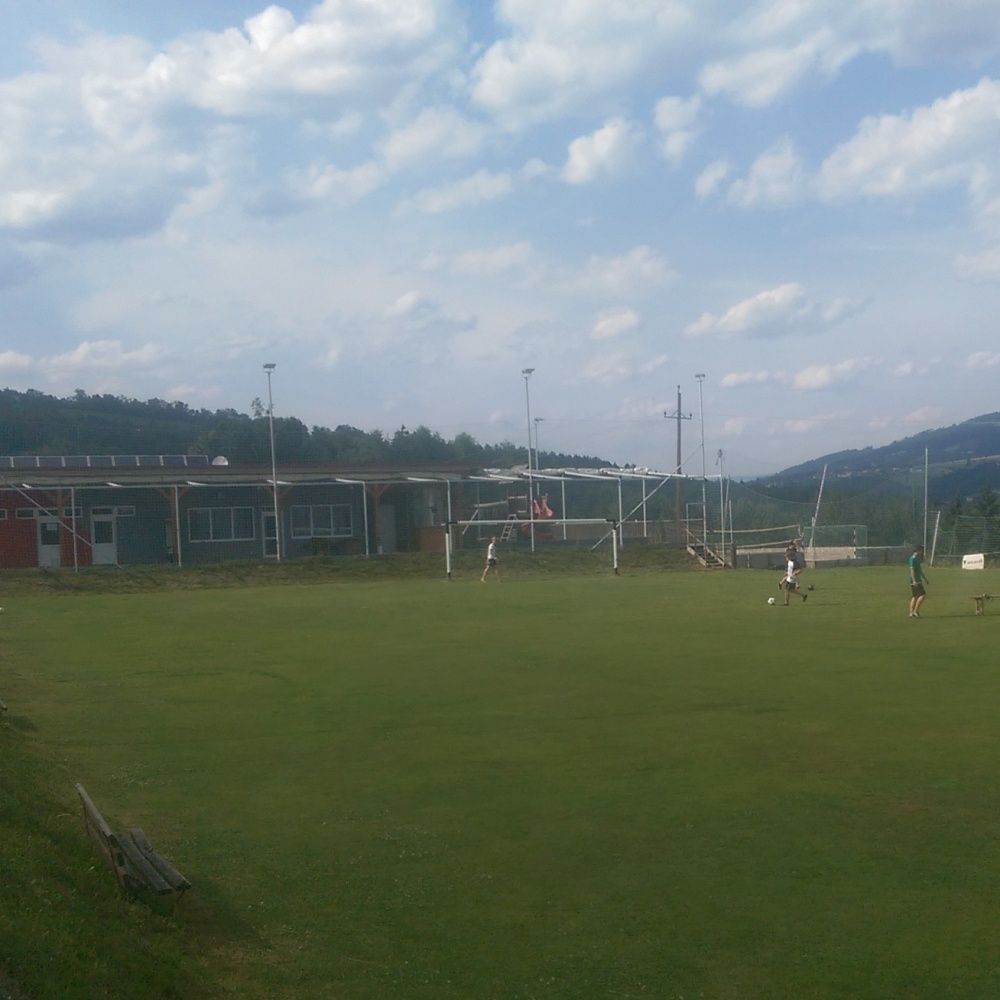 Ein Fußballfeld mit einigen Spielern unter einem teilweise bewölkten Himmel. Ein Gebäude ist im Hintergrund mit Bergen dahinter.