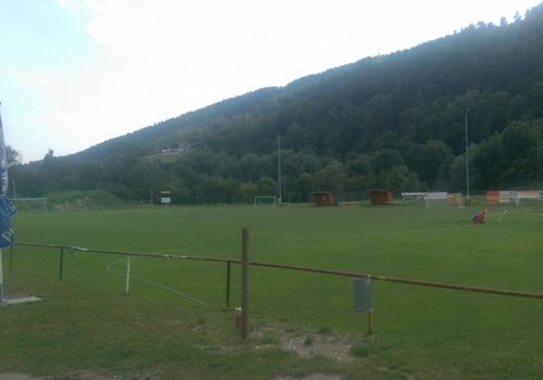 Ein Freiluft-Fußballfeld mit grünem Rasen und einem Zaun, umgeben von Bäumen und Bergen im Hintergrund. Es gibt zwei kleine Hütten auf der rechten Seite.