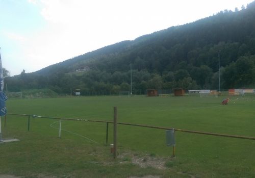 Ein Freiluft-Fußballfeld mit grünem Rasen und einem Zaun, umgeben von Bäumen und Bergen im Hintergrund. Es gibt zwei kleine Hütten auf der rechten Seite.