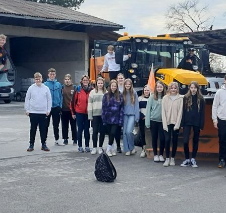 Eine Gruppe von Schülern steht vor großen gelben Baufahrzeugen in einer Garage, wobei eine Person auf einem Traktor steht.