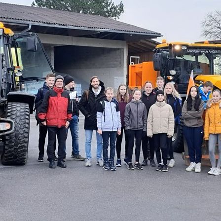 Eine Gruppe von Schülern steht vor zwei großen Maschinen. Sie alle lächeln für ein Foto. Hinter ihnen stehen zwei weitere Personen in der Nähe der Fahrzeuge.