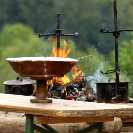 Ein Holztisch hält eine Holzschüssel mit einem Griff über einem Feuer. Zwei schwarze Töpfe mit Metallgriffen stehen auf beiden Seiten des Feuers. Der Hintergrund ist verschwommen grün.