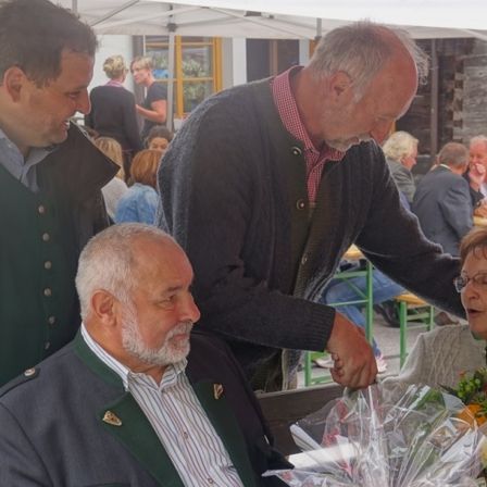 Ein Mann in einem Anzug reicht einem älteren Paar einen Blumenstrauß, das mit anderen an einem Tisch im Freien sitzt.