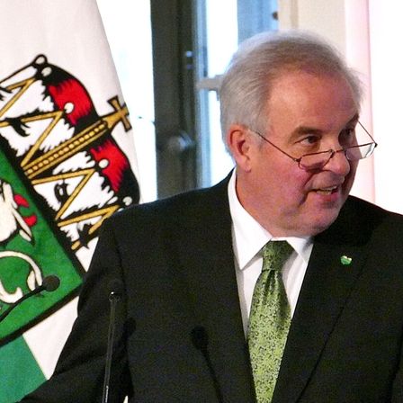 Ein älterer Mann mit grüner Krawatte und Brille steht an einem Rednerpult, mit einer Flagge im Hintergrund.