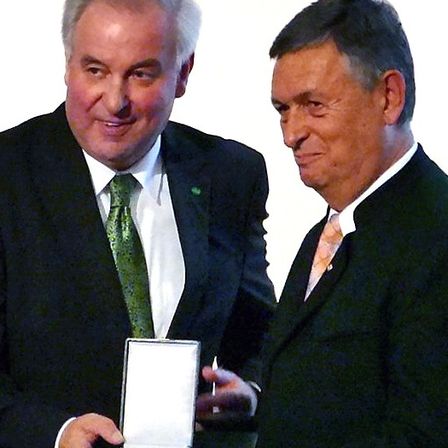 Zwei Männer in formeller Kleidung tauschen eine Auszeichnung aus. Der Mann auf der linken Seite lächelt und hält eine Schachtel. Der Mann auf der rechten Seite schaut auf die Schachtel.