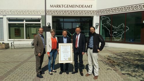 Fünf Personen stehen vor einem Gebäude mit der Aufschrift Marktgemeindeamt. Ein Mann hält ein gerahmtes Zertifikat mit 'Zertifikat' und 'Goldener Boden'.