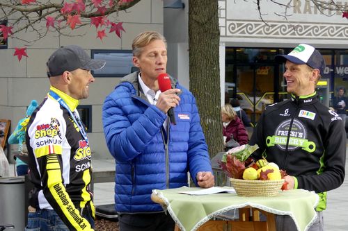 Drei Männer stehen im Freien. Der Mann in Blau hält ein Mikrofon und spricht. Hinter ihm lächelt ein Mann mit einer Kappe. Ein Mann in einem gelben und schwarzen Trikot lächelt ihn an. Ein Baum mit roten Blättern und ein Korb mit Obst stehen im Vordergrund.