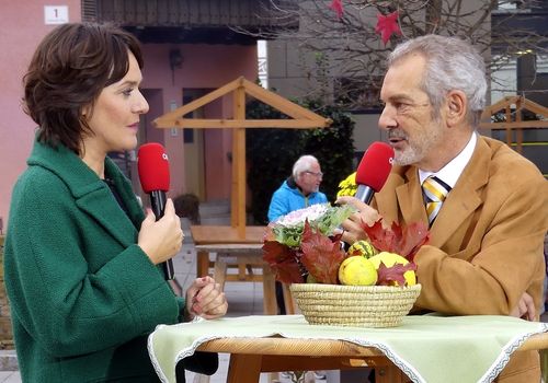 Eine Außenszene mit einer Frau in einem grünen Mantel, die ein Mikrofon hält und einen Mann in Anzug und Krawatte interviewt. Sie sitzen an einem Tisch mit einem Korb voller Früchte. Im Hintergrund geht ein Mann in einem blauen Mantel vorbei.