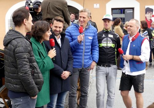 Eine Gruppe von Menschen steht im Kreis, wobei zwei Personen Mikrofone halten. Ein Mann in einem Radtrikot hält ein Mikrofon, und eine Frau in einem grünen Mantel hält ein weiteres. Sie scheinen zur Kamera zu sprechen.